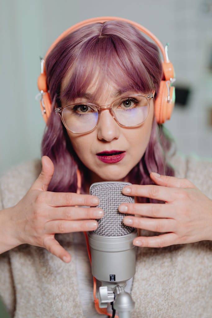 Woman with purple hair and eyeglasses using microphone for ASMR recording.